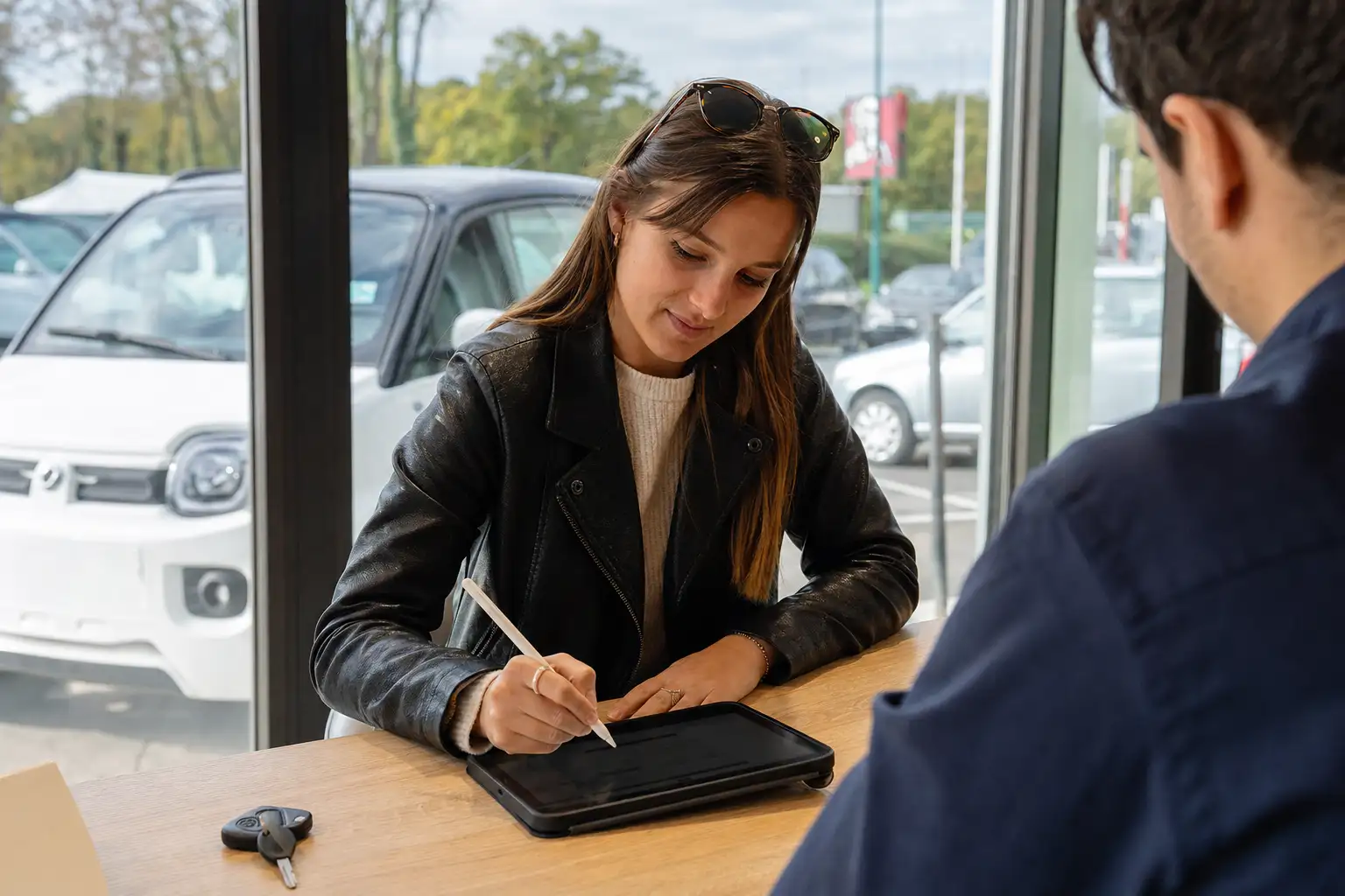 Jeune conductrice signant un contrat de location longue durée pour une voiture sans permis chez un concessionnaire, voiturette visible derrière la vitrine