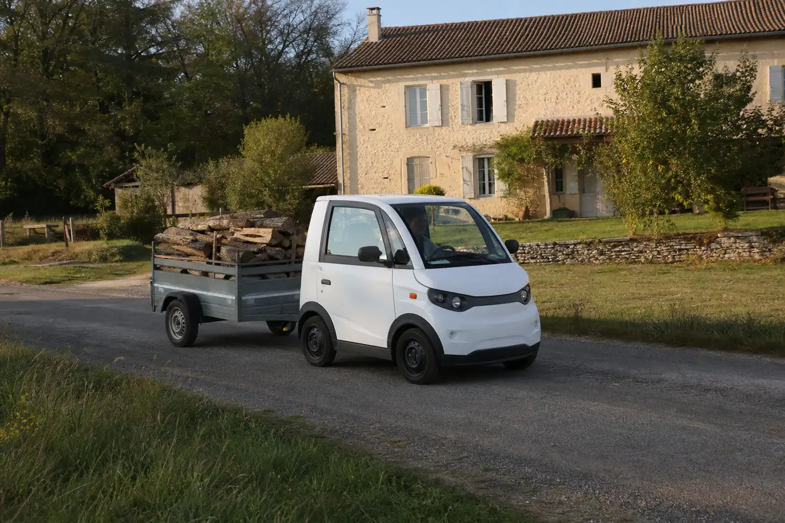 Voiture sans permis blanche tractant une petite remorque chargée de bois devant une maison de campagne française
