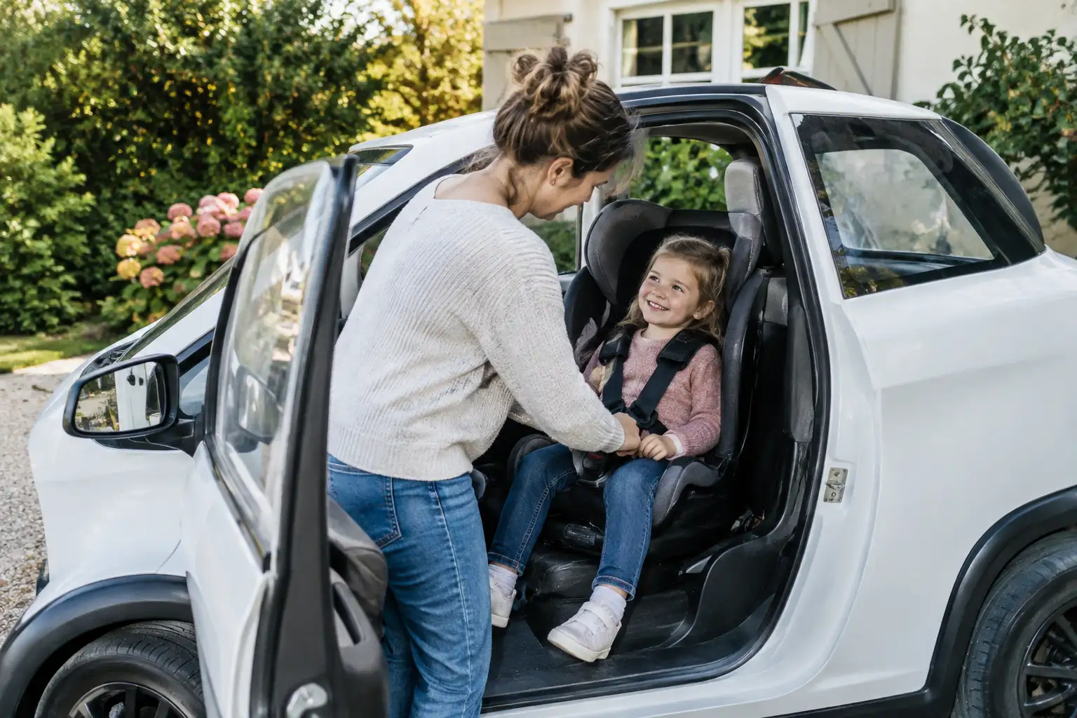 Parent installant un jeune enfant dans un siège auto sur la place passager d'une voiture sans permis garée devant une maison française